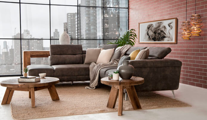 Grey L-Shape couch styled with lots of scatter cushions as well as round wooden side tables in living room with large window overlooking city scape